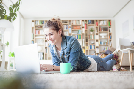 Eine junge Frau liegt in einer Wohnung auf dem Teppich und liest etwas in ihrem Laptop